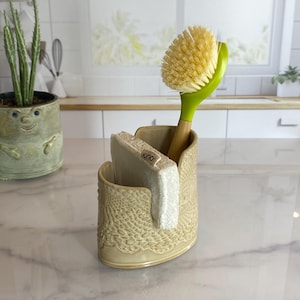 May include: A beige ceramic kitchen sink organizer with a lace-like pattern. It holds a wooden-handled dish brush with a green grip and a white sponge. A potted plant is in the background.