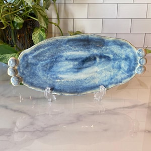 May include: A handmade, oval-shaped ceramic serving platter. The platter is glazed in shades of blue, with a marbled effect. Small, round, decorative accents are on each end. The platter is displayed on a clear stand, showcasing its unique design.