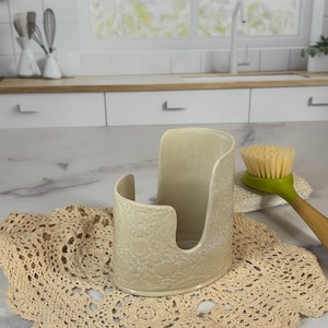 May include: A cream-colored ceramic sponge holder with a textured, lace-like pattern on the exterior. The holder features a tall, curved back and a lower front opening, designed to store a kitchen sponge upright on a countertop surface.