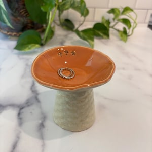 May include: A ceramic trinket dish with a glossy orange, bowl-shaped top and a textured, off-white cylindrical base. The interior of the orange bowl features three small, dark star-shaped indentations. A silver ring rests inside the dish.