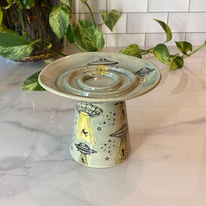 May include: A ceramic pedestal dish with a light blue glaze. The dish features a saucer-like top with concentric rings and a cylindrical base decorated with black UFOs, stars, and figures illuminated by yellow beams.