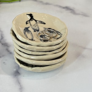 May include: A stack of five small, off-white ceramic dishes with uneven, wavy edges. The top dish features black line-art illustrations of three penguins. Two silver-colored metal rings rest inside the top dish, centered on the penguin designs.