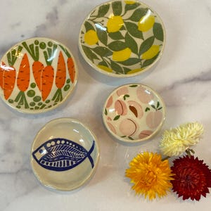 May include: A collection of small, round ceramic dishes with colorful designs. One dish features orange carrots, another lemons and green leaves, a third peaches, and the last a blue fish. Dried yellow and red flowers are also present.