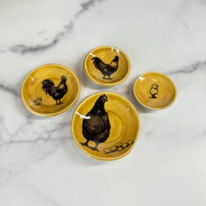 May include: A set of four round, mustard-yellow ceramic dishes featuring black ink-style illustrations of poultry. The designs include a large hen with eggs, a rooster with a small chick, a single rooster, and a small chick, all with a speckled finish.
