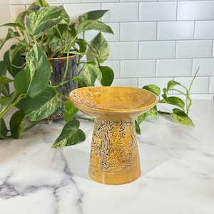 May include: A mustard yellow ceramic pedestal bowl featuring a relief design of evergreen trees and a bear. The bowl has a wide, flared rim and a cylindrical base with a glossy finish, showcasing detailed textures of a forest landscape.