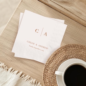 Op de afbeelding: Twee witte papieren servetten met de initialen "C | A" en de namen "Chloe & Andrew" in bruin gedrukt. De datum "Tuesday, September 2, 2029" staat ook op de servetten. Een kopje koffie staat op een geweven mat.
