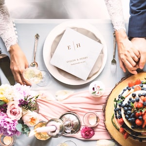 Op de afbeelding: Een bruilofttafel met een witte servet met de initialen "E H" en de namen "Evelyn & Henry" en de datum. De tafel is versierd met bloemen, kaarsen, een taart en een roze loper. Twee mensen houden elkaars handen vast.