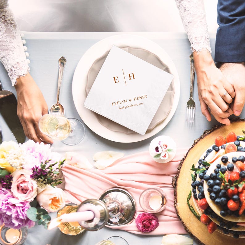 Op de afbeelding: Een bruilofttafel met een witte servet met de initialen "E H" en de namen "Evelyn & Henry" en de datum. De tafel is versierd met bloemen, kaarsen, een taart en een roze loper. Twee mensen houden elkaars handen vast.