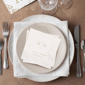 Op de afbeelding: Een tafelsetting met een witte servet en twee gepersonaliseerde servetten met de initialen "E C" en de namen "Ella & Charlie" in goud. De setting bevat een wit bord, bestek en een glas.