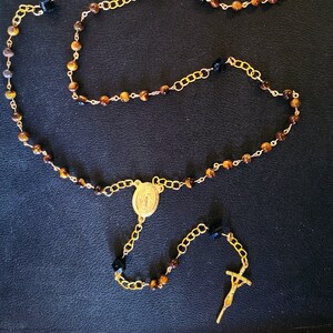 May include: A gold rosary with tiger's eye beads and black accents. The rosary features a gold pendant with a religious image and a gold cross.
