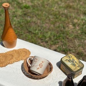May include: A collection of vintage items on a white tablecloth, including a wooden bottle, woven coasters, a floral-decorated mug in a woven basket, a small tin, and two pear-shaped salt and pepper shakers.