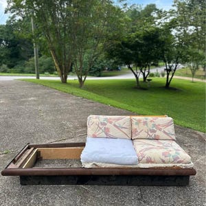 May include: A low-profile, two-seater sofa with a wooden frame and floral patterned cushions. The sofa has a dark brown base and a light blue cushion. The sofa is outdoors on a concrete surface.