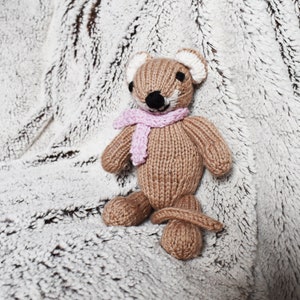 May include: A brown knitted stuffed animal with a pink scarf. The animal has a long tail and large ears. It is sitting on a white fluffy blanket.