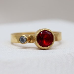 May include: A gold ring with a red gemstone and a smaller blue gemstone. The ring has a hammered texture.