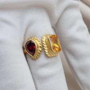 May include: Two gold rings with gemstone settings. One ring features a pear-shaped, deep red gemstone, while the other has a rectangular, amber-colored stone. Both rings have a textured, rope-like border.