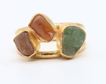 May include: A gold-colored ring featuring three irregularly shaped gemstones in shades of brown, orange, and green. The ring has a unique design with an open space between the gemstones, set against a white background.