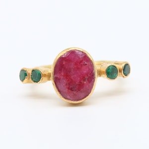 May include: A gold ring with a large, red, oval-shaped ruby gemstone surrounded by smaller, green gemstones.