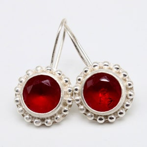 Op de afbeelding: Een paar zilveren oorbellen met rode edelsteen accenten. De oorbellen zijn rond van vorm met een parelrand.