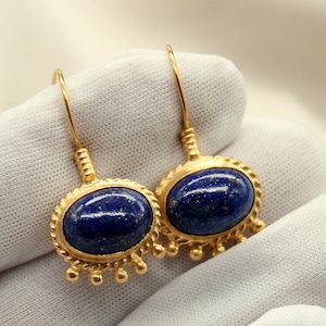May include: A pair of gold earrings featuring oval, deep blue gemstones. The earrings have a decorative border and small, dangling accents. The jewellery is displayed on a white surface.