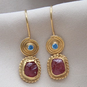 Op de afbeelding: Gouden oorbellen met een rode edelsteen en een blauwe edelsteen. De oorbellen hebben een spiraalvormig ontwerp.