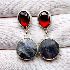 May include: A pair of silver earrings with red and blue gemstone accents. The top of each earring features a red oval-shaped gemstone, and the bottom features a round blue gemstone.