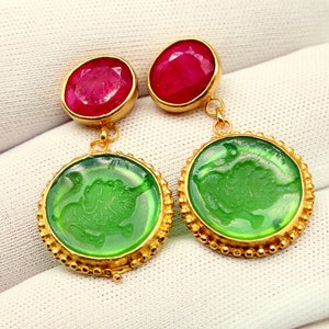 Op de afbeelding: Een paar gouden oorbellen met groene glazen druppels met een gesneden ontwerp en rode edelsteenaccenten.