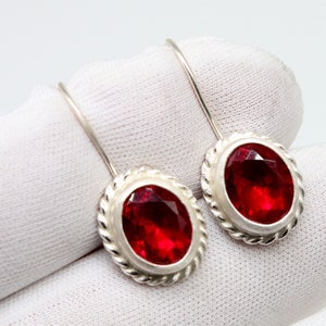 Peut inclure: Une paire de boucles d'oreilles en argent avec des pierres précieuses ovales rouges. Les boucles d'oreilles ont un design simple avec une bordure en forme de corde autour des pierres précieuses.
