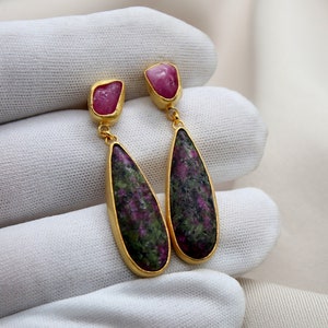 Op de afbeelding: Gouden oorbellen met een traanvormige groene en roze edelsteen. De oorbellen hebben een kleine roze edelsteen aan de bovenkant en een grotere traanvormige edelsteen eronder.