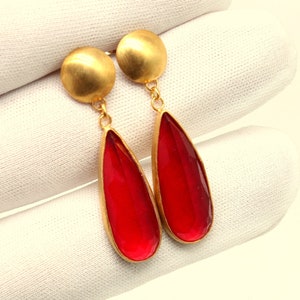 Op de afbeelding: Een paar gouden oorhangers met rode traanvormige stenen.