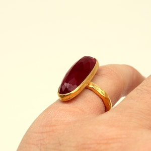 Może przedstawiać: Złoty pierścionek z dużym, owalnym, ciemnoczerwonym kamieniem szlachetnym.