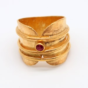 May include: A gold ring with a textured, hammered finish and a single red gemstone in the center. The ring is wide and has a unique, organic shape.