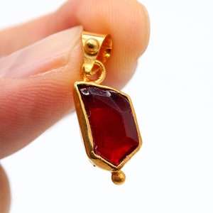 Pode incluir: Um pingente de ouro com uma gema vermelha. O pingente tem a forma de um diamante e tem uma pequena bola de ouro na parte inferior.