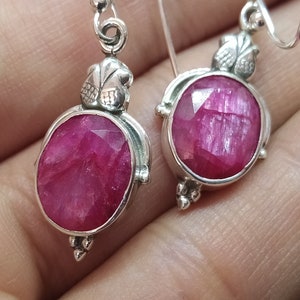 Op de afbeelding: Een paar zilveren oorbellen met roze robijn edelstenen. De oorbellen hebben een bloemmotief en zijn ovaalvormig.