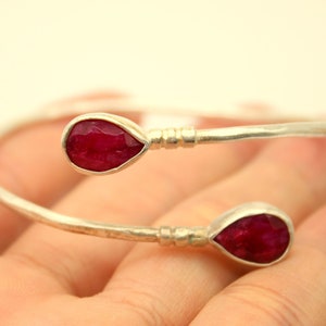May include: A silver bangle bracelet with two teardrop-shaped ruby gemstones. The bracelet is open and has a hammered finish.