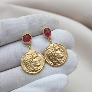 Può includere: Orecchini d'oro con una pietra preziosa rossa e un ciondolo a forma di moneta che presenta il profilo di un viso.