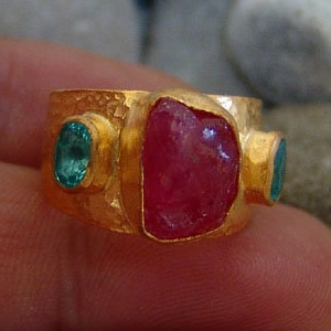 Pode incluir: Um anel de ouro com uma grande pedra vermelha e duas pedras verdes menores. O anel é engastado em uma banda texturizada.