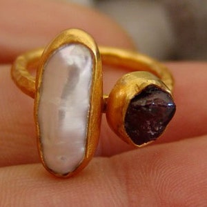 May include: Gold ring with a white pearl and a red gemstone. The ring is on a finger.
