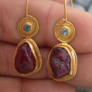 Könnte beinhalten: Ein Paar goldene Ohrringe mit einem tropfenförmigen roten Edelstein und einem kleineren blauen Edelstein. Die Ohrringe haben ein zartes Design und sind perfekt, um jedem Outfit einen Hauch von Eleganz zu verleihen.