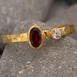 Pode incluir: Um anel de ouro com uma gema oval vermelha e uma gema redonda branca menor. O anel tem uma textura martelada.
