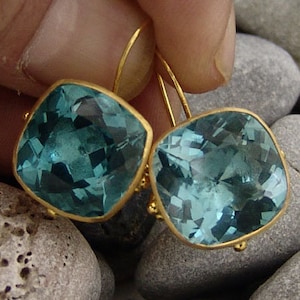 May include: A pair of gold earrings with square blue gemstones. The earrings are hanging from a person's hand.