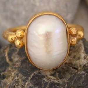 Könnte beinhalten: Ein goldener Ring mit einem weißen Perlmutt, das in der Mitte eingesetzt ist. Der Ring hat eine strukturierte Band mit kleinen goldenen Perlen auf beiden Seiten des Perlmutts.