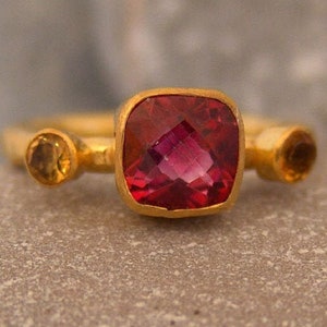 May include: A gold ring with a large, square, red gemstone and two smaller, round, yellow gemstones.