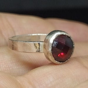 May include: A silver ring featuring a faceted, deep red gemstone. The round gemstone is set in a bezel setting, and the band is a simple, flat design. The ring is shown against a neutral background.