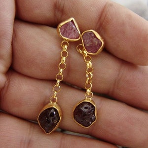 May include: A pair of gold earrings with pink and dark red gemstone drops. The earrings feature a delicate chain design.