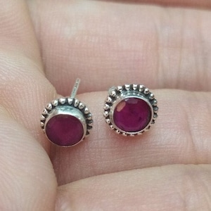 Op de afbeelding: Een paar zilveren oorbelletjes met een ronde, donkerroze edelsteen die in elk is gezet. De oorbelletjes hebben een parelrand rond de edelsteen.
