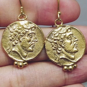 Op de afbeelding: Gouden oorbellen met een rond, muntvormig ontwerp met een profiel van een mannengezicht. De oorbellen hebben een eenvoudige haaksluiting.