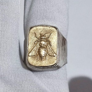 Op de afbeelding: Een zilveren ring met een rechthoekig gouden bijenontwerp.