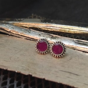 May include: A pair of silver stud earrings with a small, round, dark pink gemstone in the center of each earring.