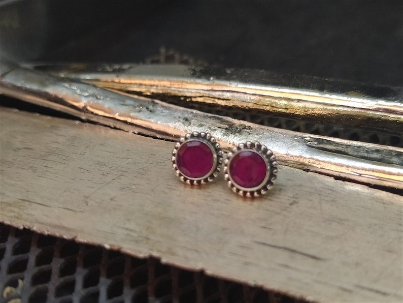 May include: A pair of silver stud earrings with a small, round, dark pink gemstone in the center of each earring.