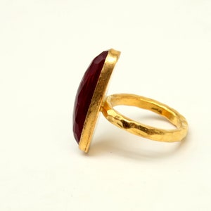 Może przedstawiać: Złoty pierścionek z dużym, fasetowanym, ciemnoczerwonym kamieniem szlachetnym. Obrączka pierścionka jest fakturowana i ma młotkowane wykończenie.